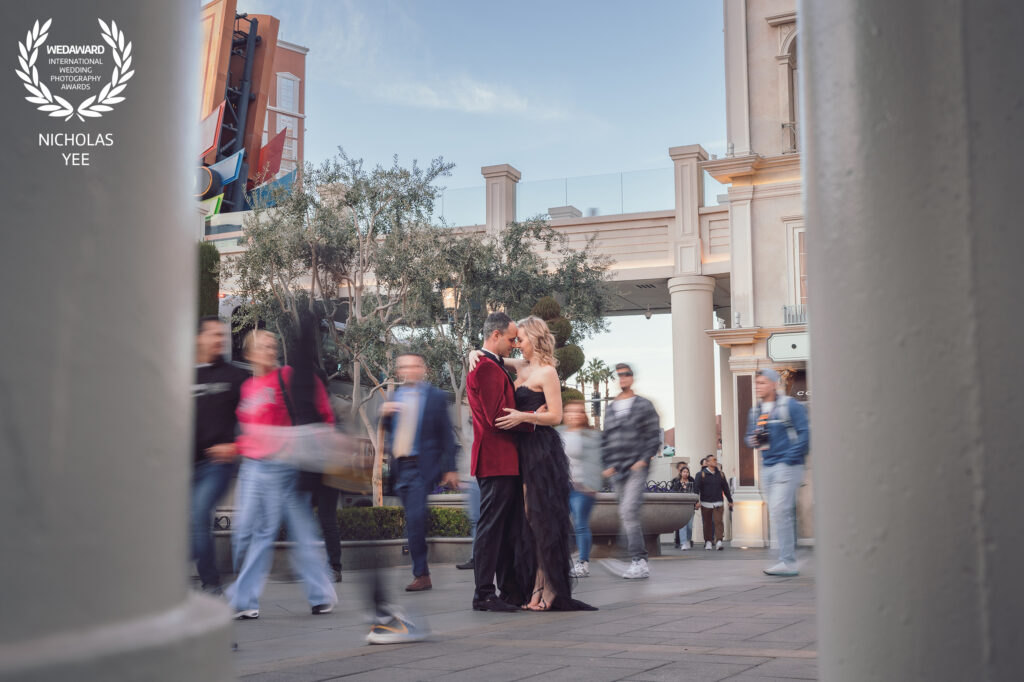 Couple embracing in the middle of a busy urban plaza with blurred pedestrians walking by, the groom in a red jacket and the bride in a black dress, featured in the WedAward Collection #91.