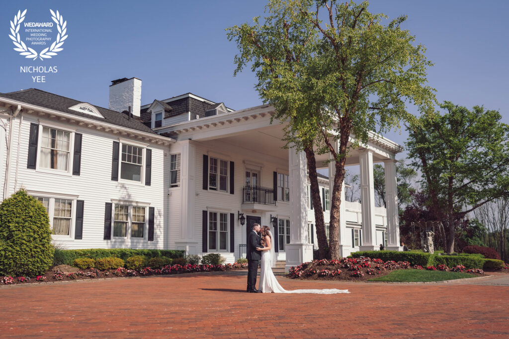Couple standing in front of a stately white mansion with columns, the bride’s long veil trailing on the red brick driveway, featured in the WedAward Collection #94.