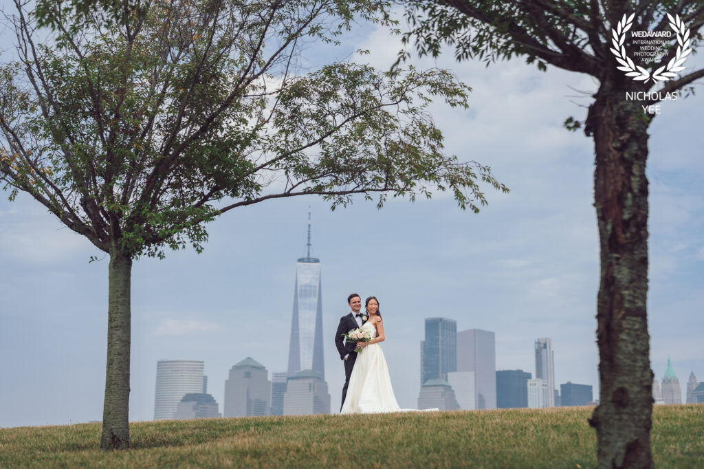 Bride and groom posing on a grassy hill framed by two trees, with the New York City skyline, including the One World Trade Center, in the background, included in the WedAward Collection #94.