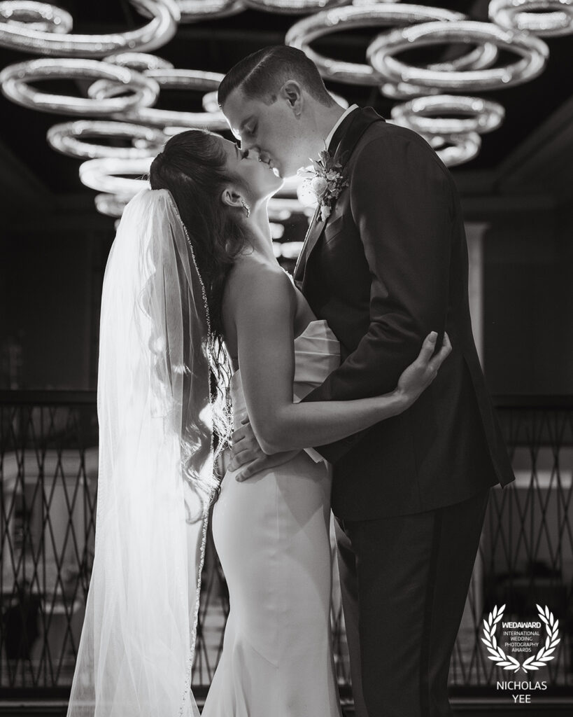 Black and white photo of a bride and groom sharing a kiss indoors under modern circular light fixtures, with the bride’s veil flowing behind her, part of the WedAward Collection #100.