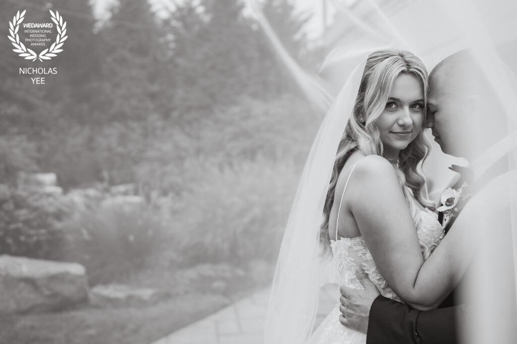 Bride looking directly at the camera while embracing the groom outdoors, both wrapped in the bride’s veil, featured in the WedAward Collection #100.