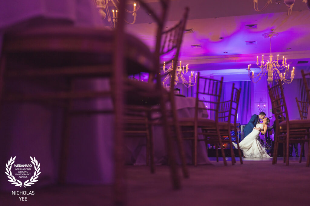Bride and groom sharing a romantic dip kiss on the dance floor of an elegantly lit ballroom with purple lighting and chandeliers, part of the WedAward Collection #102.