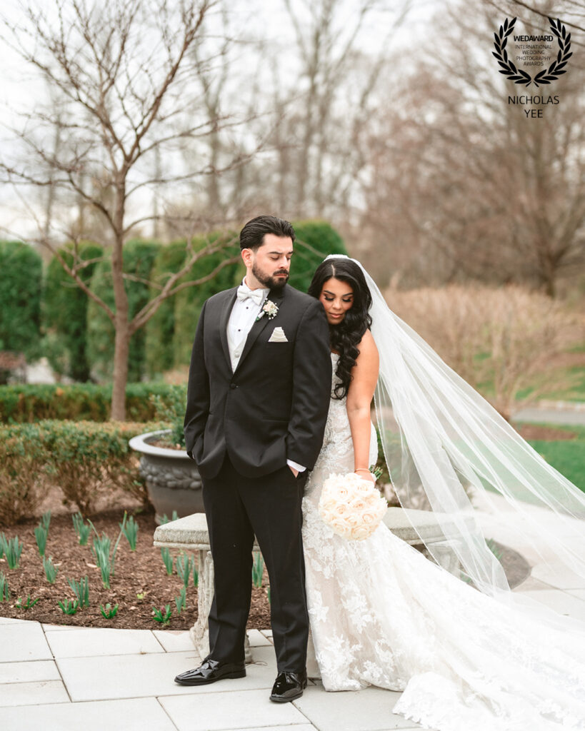 Couple posing outdoors on a stone path, with the bride’s long veil flowing behind her, both dressed elegantly for their wedding day, included in the WedAward Collection #103.
