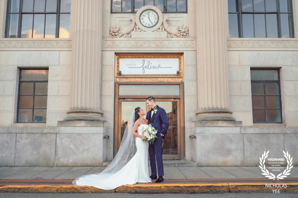 Bride and groom standing close and smiling in front of a building with a clock and a sign reading “Felina,” with the bride holding a white bouquet, captured in the WedAward Collection #103.
