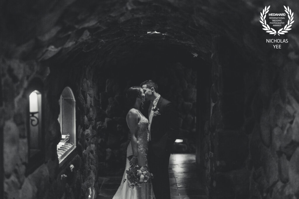 Black and white photo of bride and groom sharing a kiss inside a dimly lit stone archway, creating an intimate and timeless moment, featured in the WedAward Collection #103.