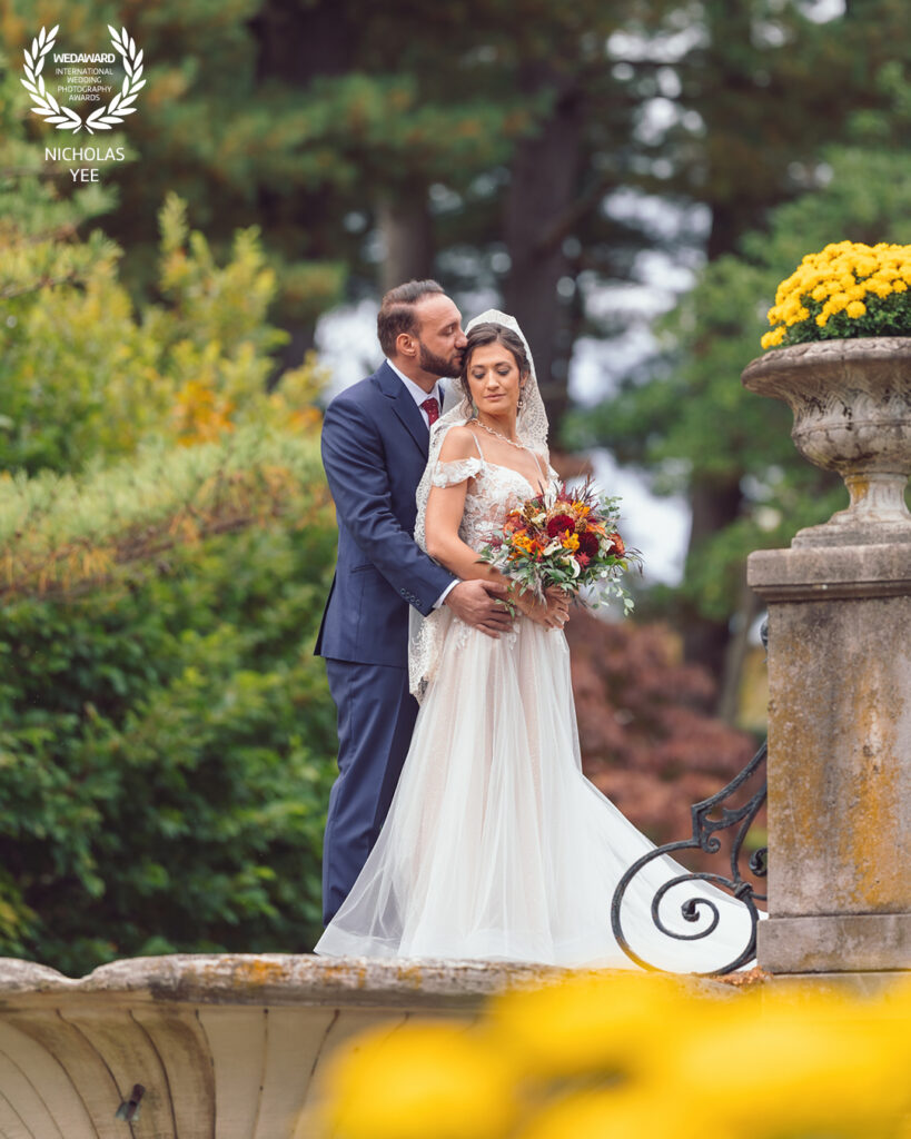 Groom tenderly kissing bride’s temple as she holds a vibrant bouquet, standing outdoors surrounded by lush greenery and yellow flowers, part of the WedAward Collection #103.