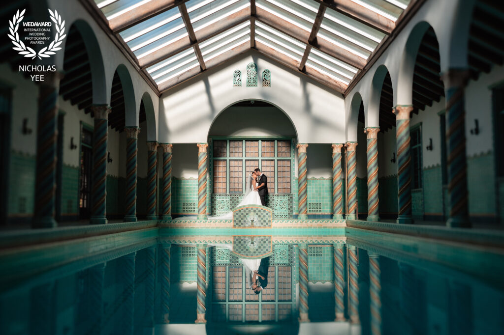 Bride and groom embracing in an ornate indoor pool area with detailed tile work and tall arched windows, their reflection visible in the water, part of the WedAward Collection #104.