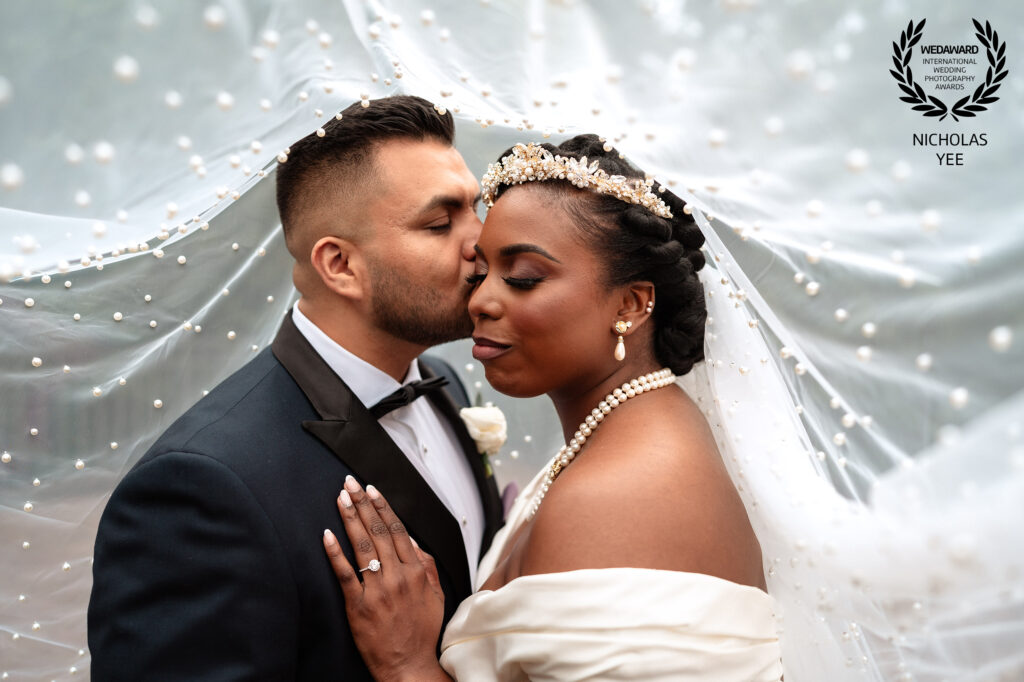 Groom gently kissing bride’s cheek as she closes her eyes, both dressed elegantly with the bride’s pearl-adorned veil flowing behind them, featured in the WedAward Collection #104.