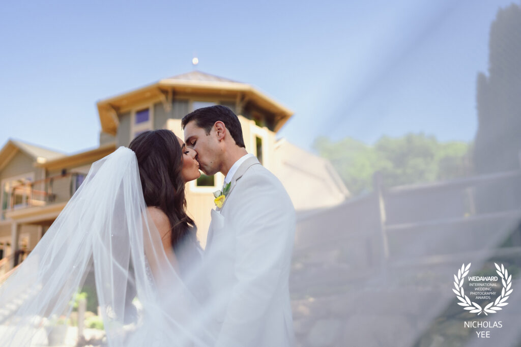 Bride and groom sharing a tender kiss outside a rustic building, with the bride’s veil softly framing the scene, captured in the WedAward Collection #105.