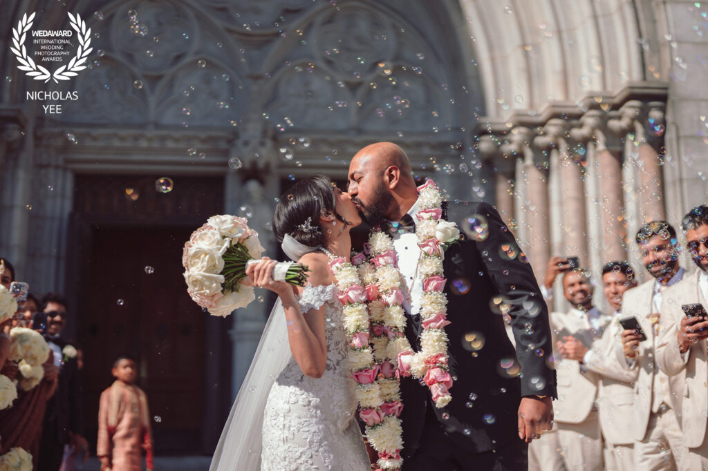 Newlywed couple kissing amid bubbles and celebratory guests outside a grand church, the bride holding a bouquet and both wearing floral garlands, featured in the WedAward Collection #105.