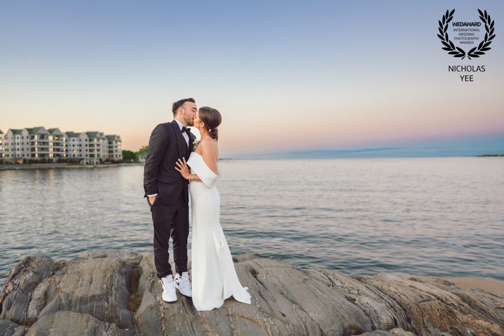 Bride and groom sharing a kiss on rocky shore during sunset with calm ocean and buildings in the background, featured in the WedAward Collection.
