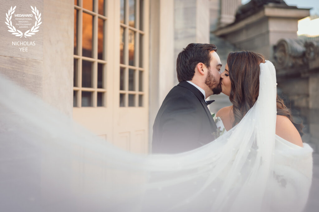 Newlywed couple kissing with the bride’s veil flowing dramatically in front of a classic building, featured in the WedAward Collection #90.