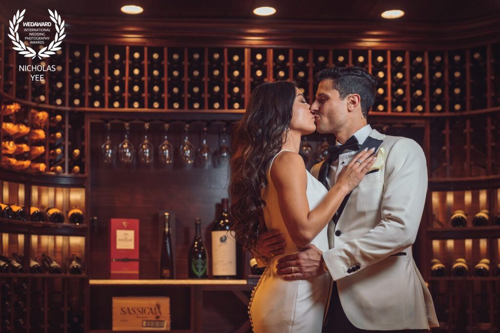 Couple embracing and about to kiss in a cozy wine cellar surrounded by shelves of wine bottles, featured in the WedAward Collection #92.
