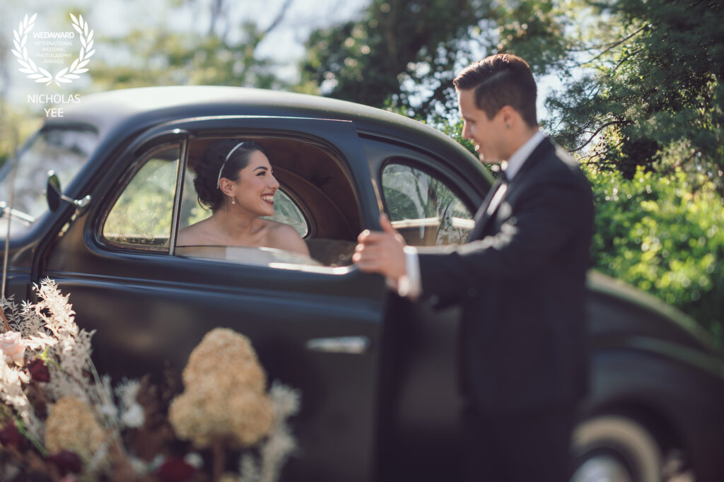 Bride smiling joyfully from inside a vintage black car as the groom opens the door for her outdoors, included in the WedAward Collection #92.