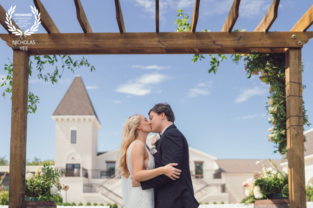 Bride and groom kissing under a wooden pergola with a church building and blue sky in the background, part of the WedAward Collection #96.