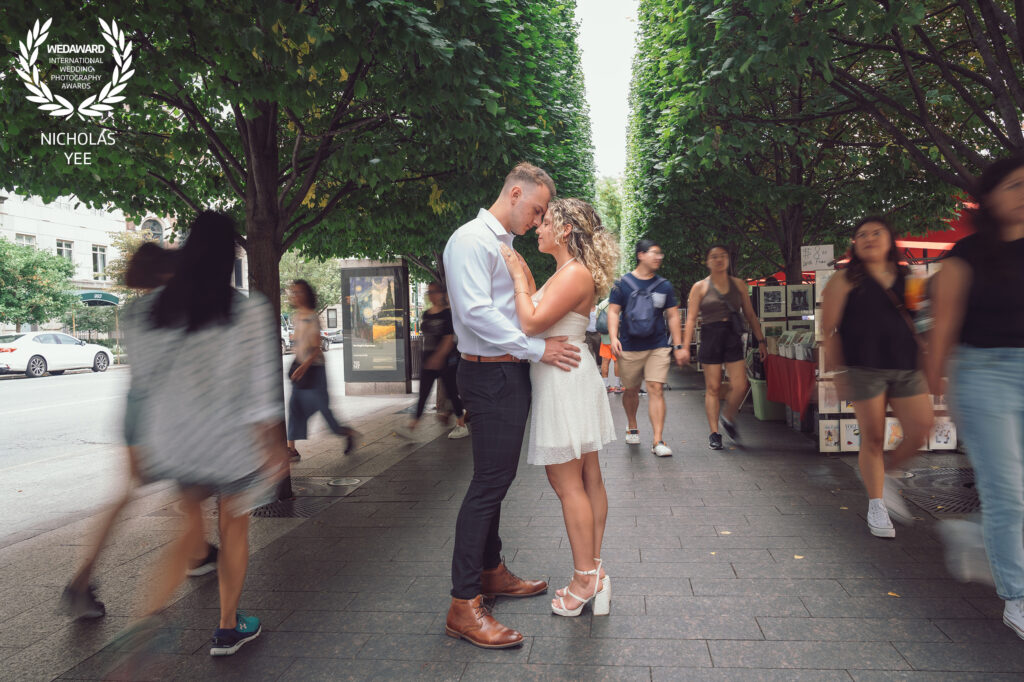 Couple embracing and kissing in the middle of a busy urban street lined with trees, surrounded by passersby, featured in the WedAward Collection #96.