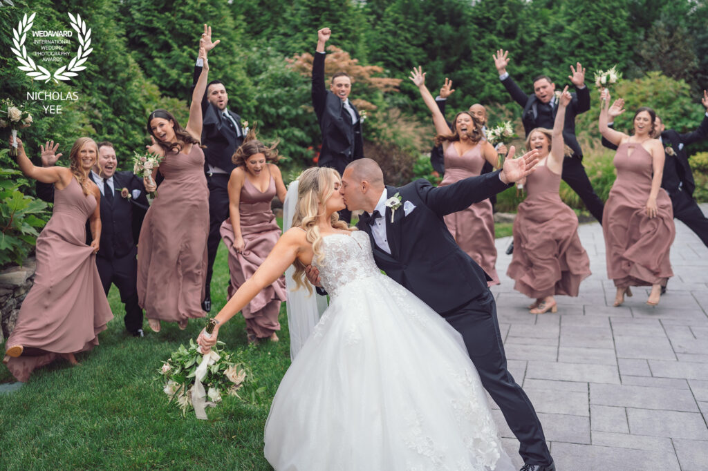 Bride and groom sharing a romantic kiss while the bridal party cheers joyfully behind them outdoors, with bridesmaids in mauve dresses and groomsmen in black suits, part of the WedAward Collection #97.