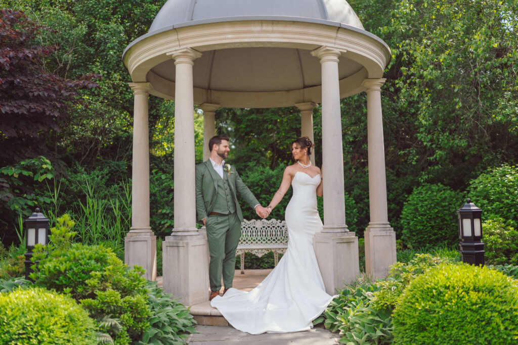 Bride and groom holding hands under a gazebo during their wedding day, captured as part of a wedding day timeline, showcasing an intimate moment in a beautiful garden setting.