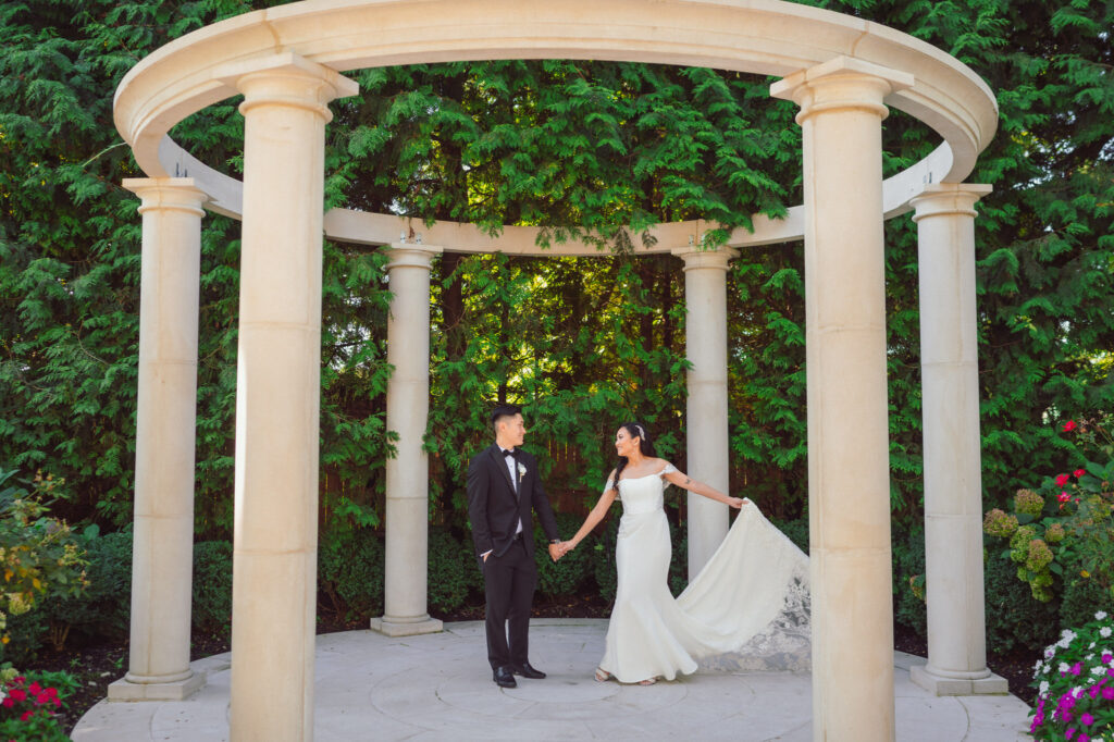 Couple holding hands under a stone gazebo, with the bride's gown flowing elegantly, captured during their wedding day timeline, highlighting a romantic and scenic moment.