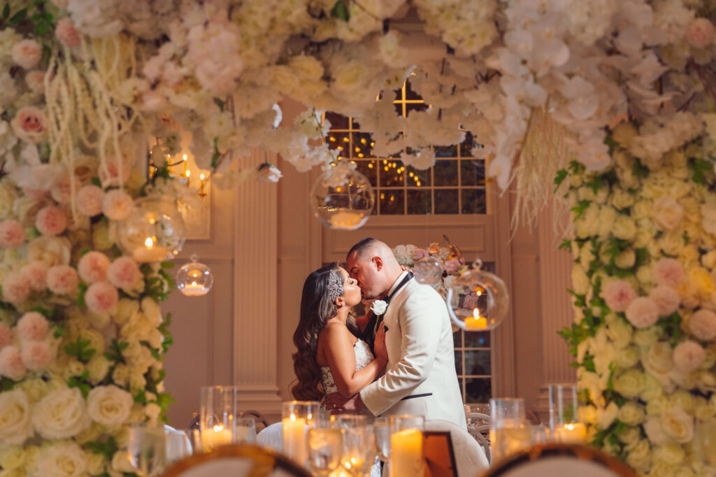 A bride and groom share a romantic kiss under a lush floral arch with soft candlelight and hanging glass globes, perfectly captured by Nicholas Yee Photography with no hidden fees or extra charges.