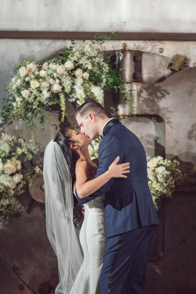 Bride and groom sharing a tender kiss inside a rustic venue adorned with white floral arrangements, the groom gently holding the bride.