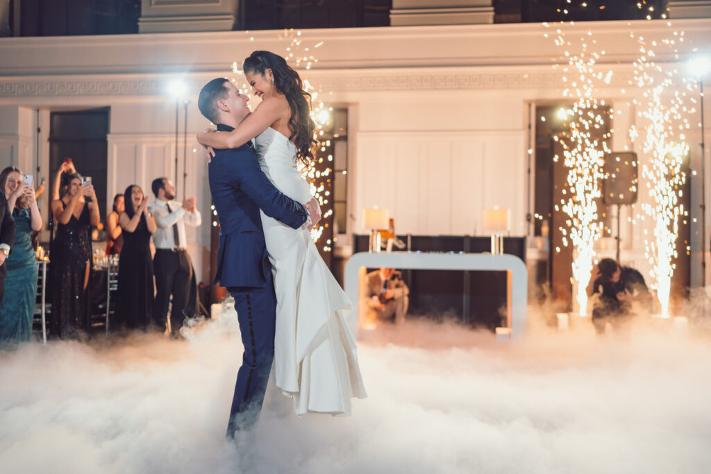 Bride and groom sharing their first dance on a fog-covered floor with sparkling pyrotechnics and joyful guests in the background, inside an elegant indoor setting.