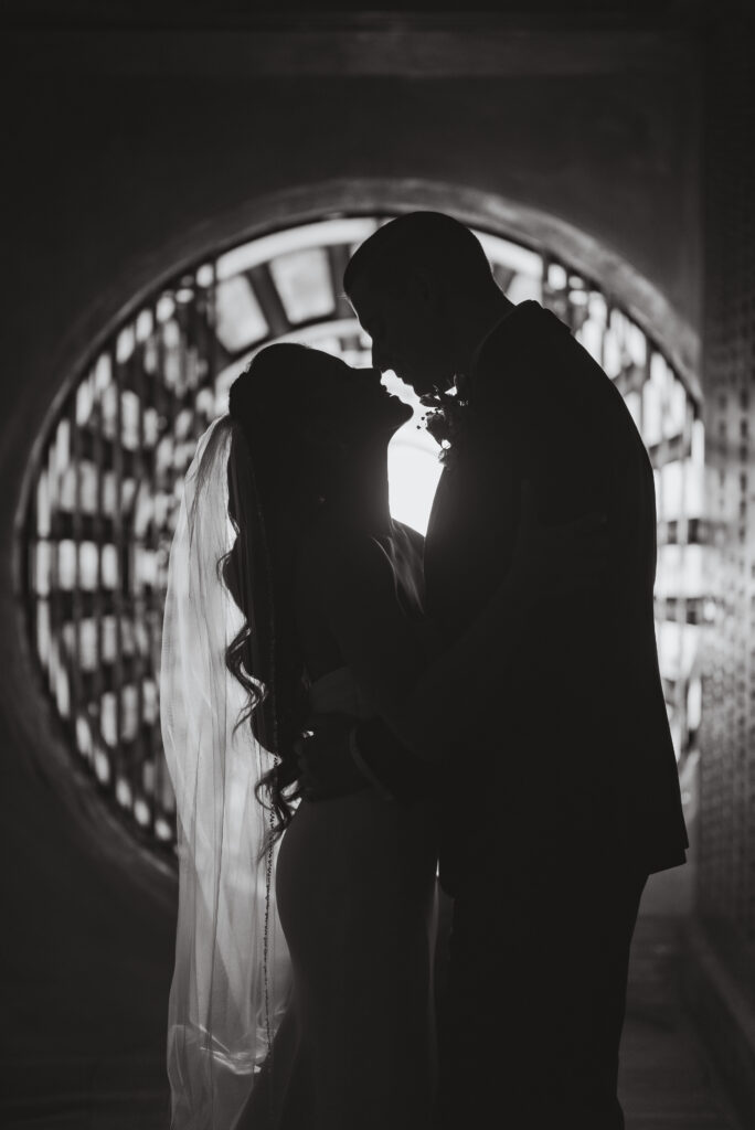 Silhouetted bride and groom in an intimate embrace inside a dimly lit, arched hallway, the bride’s veil softly catching light behind them.