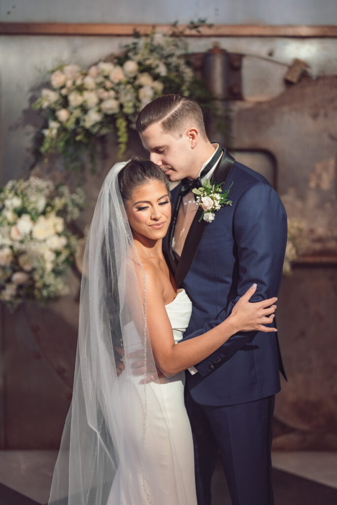 Bride and groom smiling and holding hands, surrounded by greenery and flowers, the bride in a white dress and the groom in a blue suit.