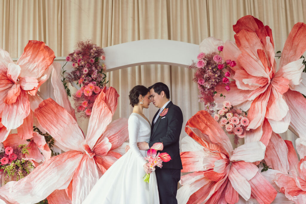 Full-length portrait of a bride and groom standing close with foreheads touching, framed by large pink and coral paper flowers, captured elegantly by Nicholas Yee Photography without hidden fees or extra charges.