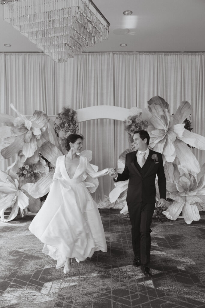 Black and white image of the bride and groom walking hand-in-hand, surrounded by oversized floral decorations, demonstrating the candid style of Nicholas Yee Photography with no hidden fees or extra charges.