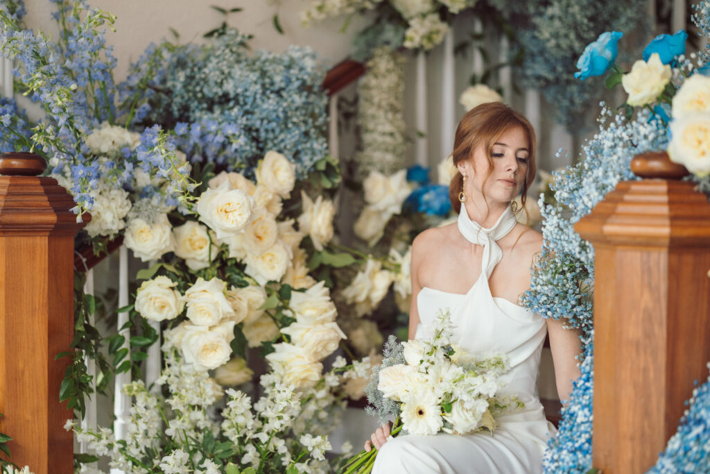 A red-haired bride in an elegant white halter-neck dress holding a bouquet of white and blue flowers, sitting thoughtfully amid lush floral arrangements, captured beautifully by Nicholas Yee Photography with no hidden fees or extra charges.
