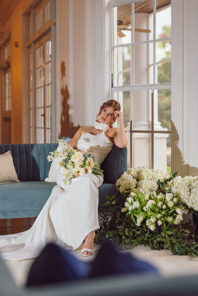 The same bride in the white dress, seated on a teal sofa near large windows, surrounded by white and blue flowers, showcasing the refined style of Nicholas Yee Photography without hidden fees or extra charges.