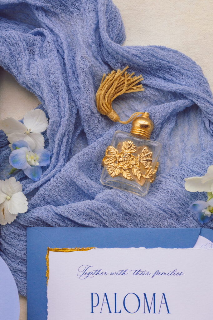 Close-up of a vintage gold-trimmed perfume bottle resting on a blue fabric with wedding invitation details, artfully styled and photographed by Nicholas Yee Photography, emphasizing no hidden fees or extra charges.