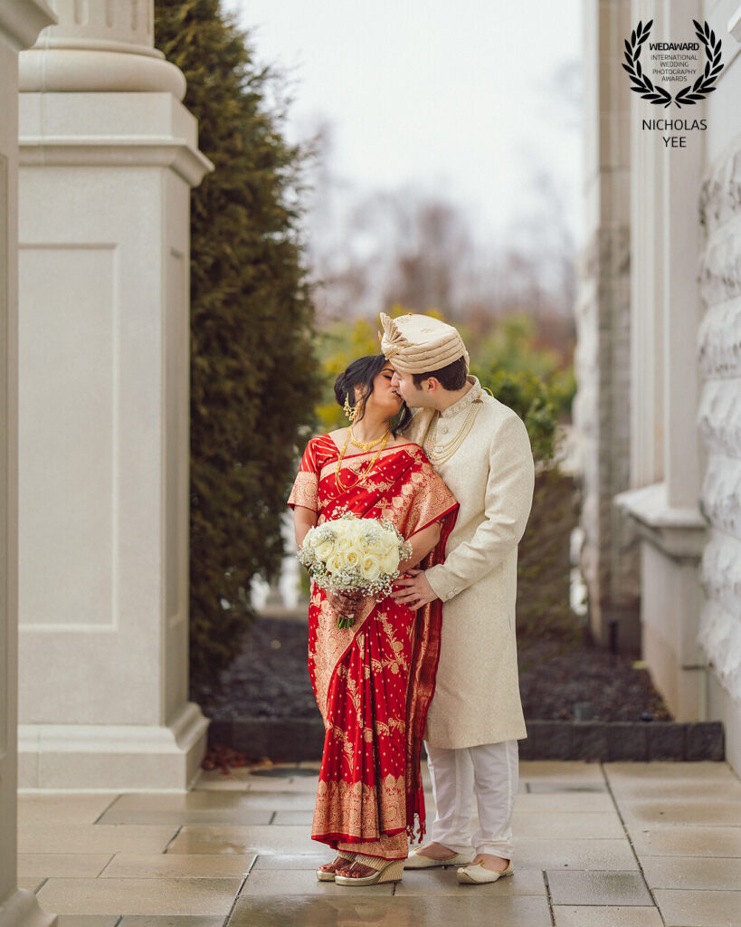 A couple dressed in traditional Indian wedding attire shares a tender kiss outdoors, with the bride wearing a red and gold saree holding a bouquet of white roses. This romantic moment is part of the WedAward Collection #116, captured by photographer Nicholas Yee.