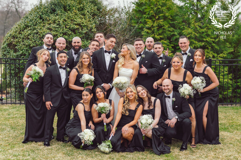 A joyful wedding party from WedAward Collection #116 featuring the bride in a white gown and groom in a tuxedo surrounded by bridesmaids in black dresses holding white floral bouquets and groomsmen in black tuxedos, posing outdoors with greenery in the background.