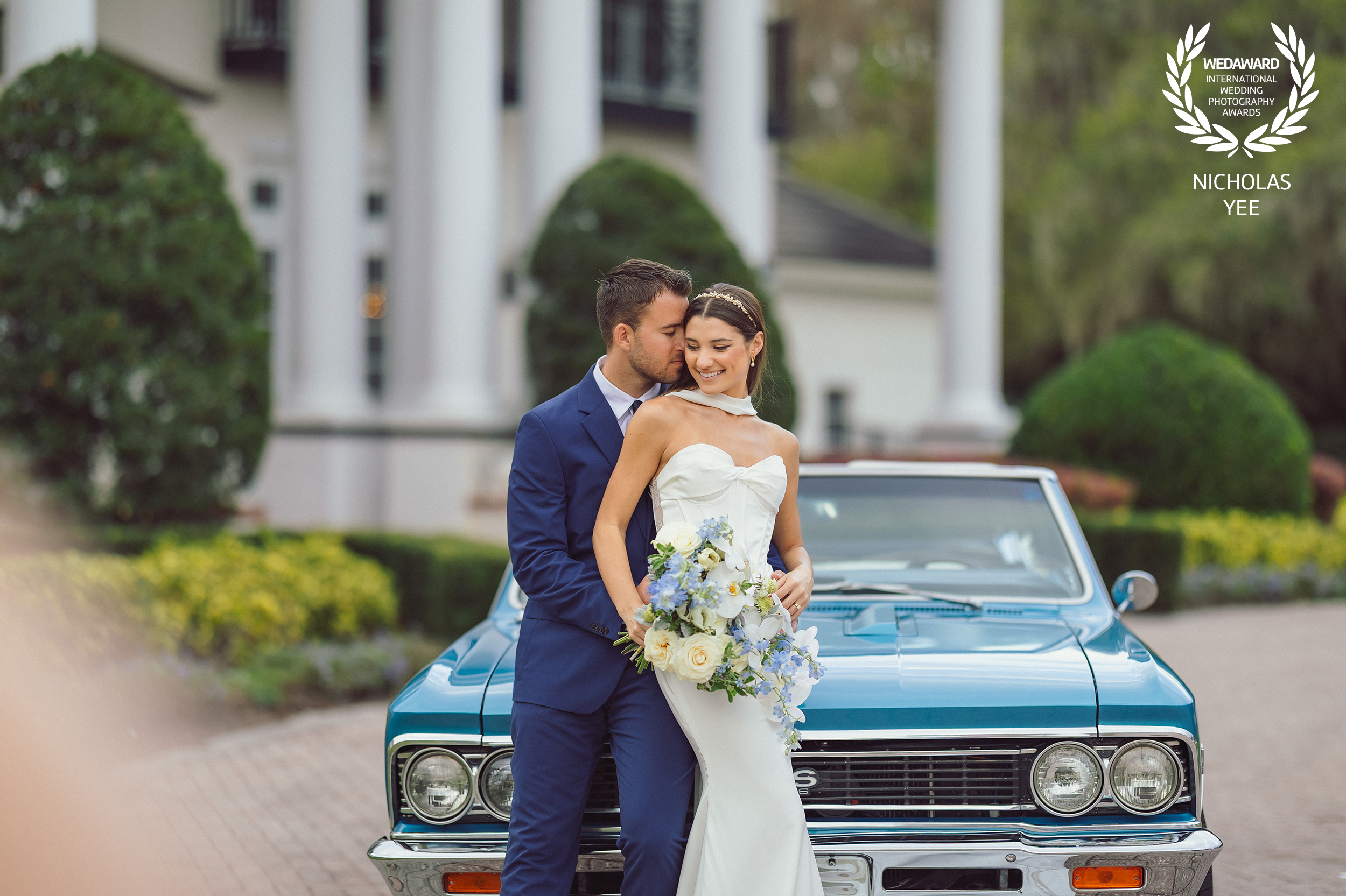 A couple standing in front of a vintage car, with the bride holding a bouquet of blue and white flowers while her partner embraces her from behind. The image is awarded as part of the WedAward Collection #115 by Nicholas Yee Photography