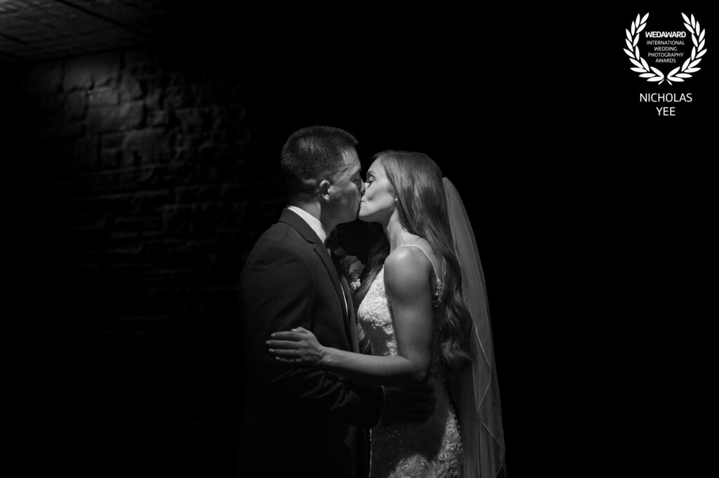 WedAward Collection #120 winning image by Nicholas Yee Photography. A romantic, black-and-white photo of a bride and groom sharing a kiss in a dimly lit space, captured with emotion and timeless elegance.
