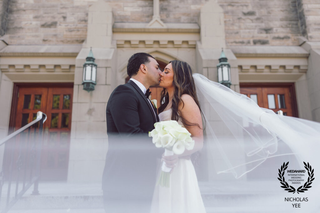 WedAward Collection #120 winning image by Nicholas Yee Photography. A romantic, black-and-white photo of a bride and groom sharing a kiss in a dimly lit space, captured with emotion and timeless elegance.