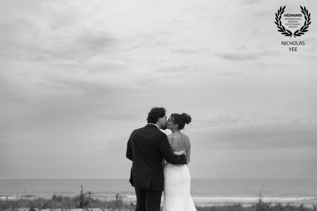 A couple kissing on the beach with the ocean and cloudy sky in the background, captured in black and white. A featured photo in WedAward Collection #124.