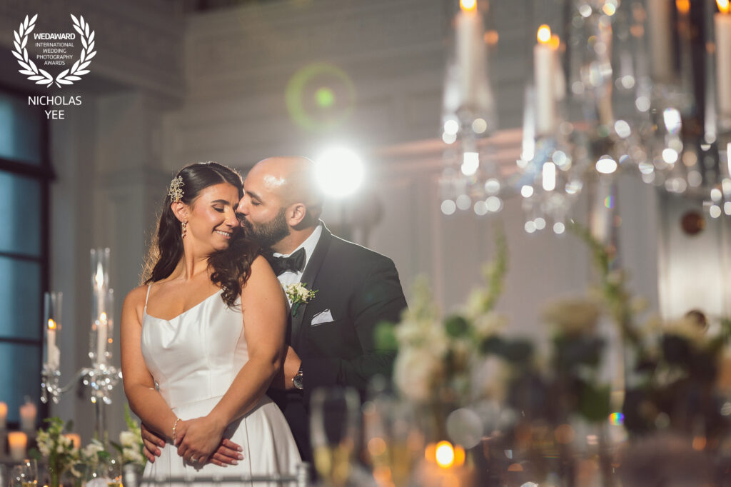 Couple embraces in a romantic moment during their wedding reception, captured in an elegant, soft-lit setting with candles and chandeliers. Award-winning wedding photography by Nicholas Yee, capturing real, intimate connections.