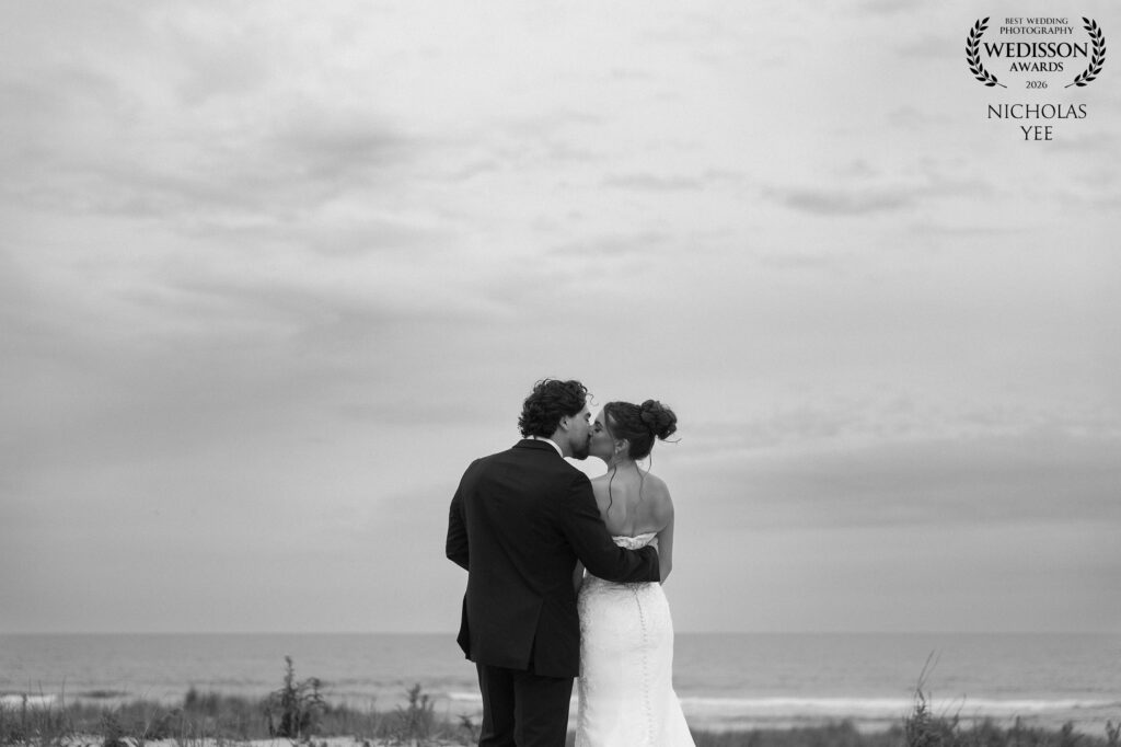 Wedisson Collection #130 Winner: A couple shares a tender kiss on the beach, surrounded by the vast ocean and soft, cloudy skies. The intimate moment is captured in black-and-white, evoking timelessness and deep emotional connection