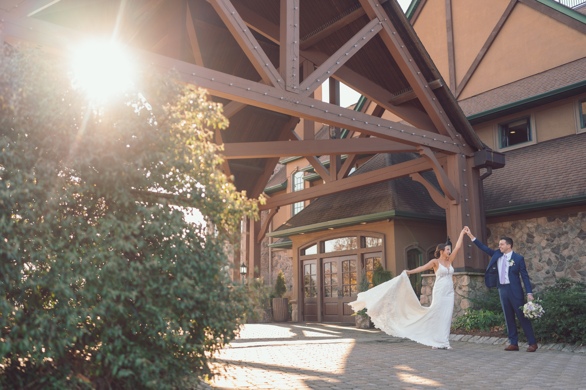 Bride and groom dancing joyfully in front of a beautifully rustic wedding venue with warm sunlight filtering through the trees.