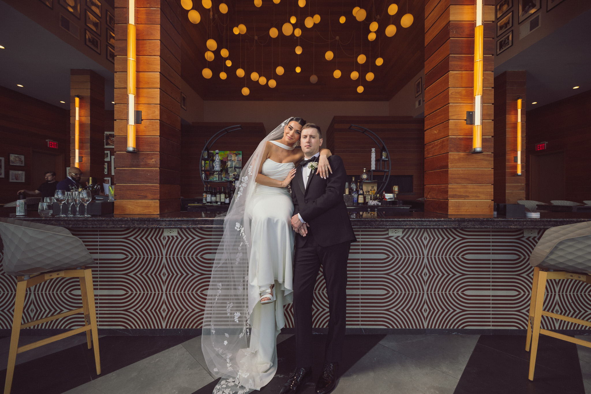 Bride and groom posing at the bar in Hudson House, Jersey City, NJ. The bride, wearing a sleek white gown and long veil, sits on the bar counter while the groom stands beside her in a classic black tuxedo. The warm ambiance is highlighted by stylish lighting and modern décor in the background.