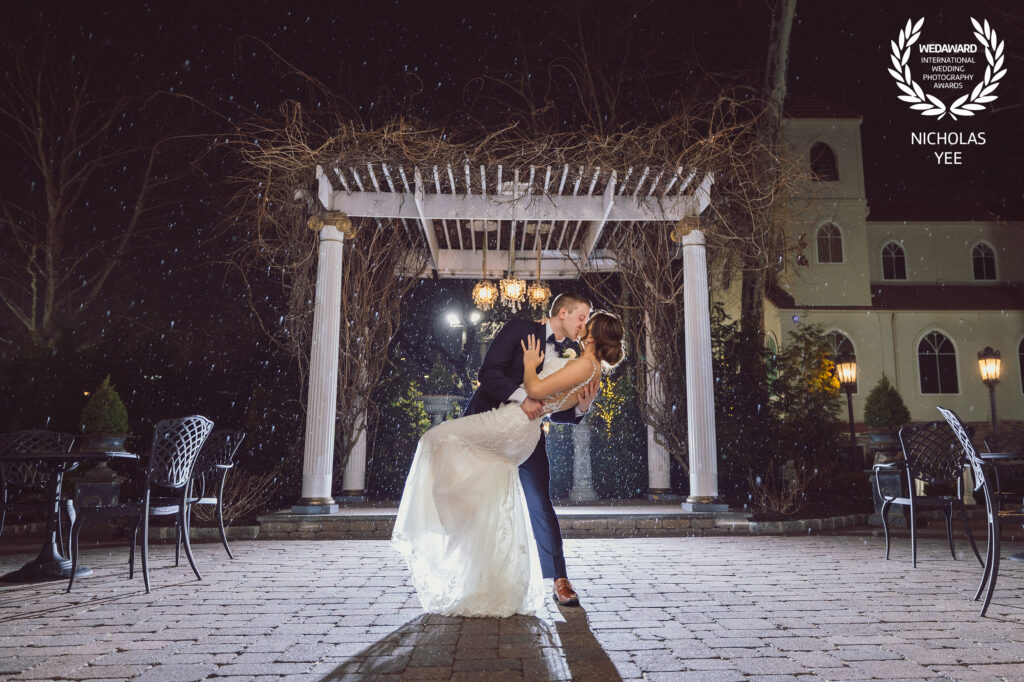 A beautifully lit kiss captured under a charming wedding arbor with snow falling, showcasing the artistry of an award-winning wedding photographer.