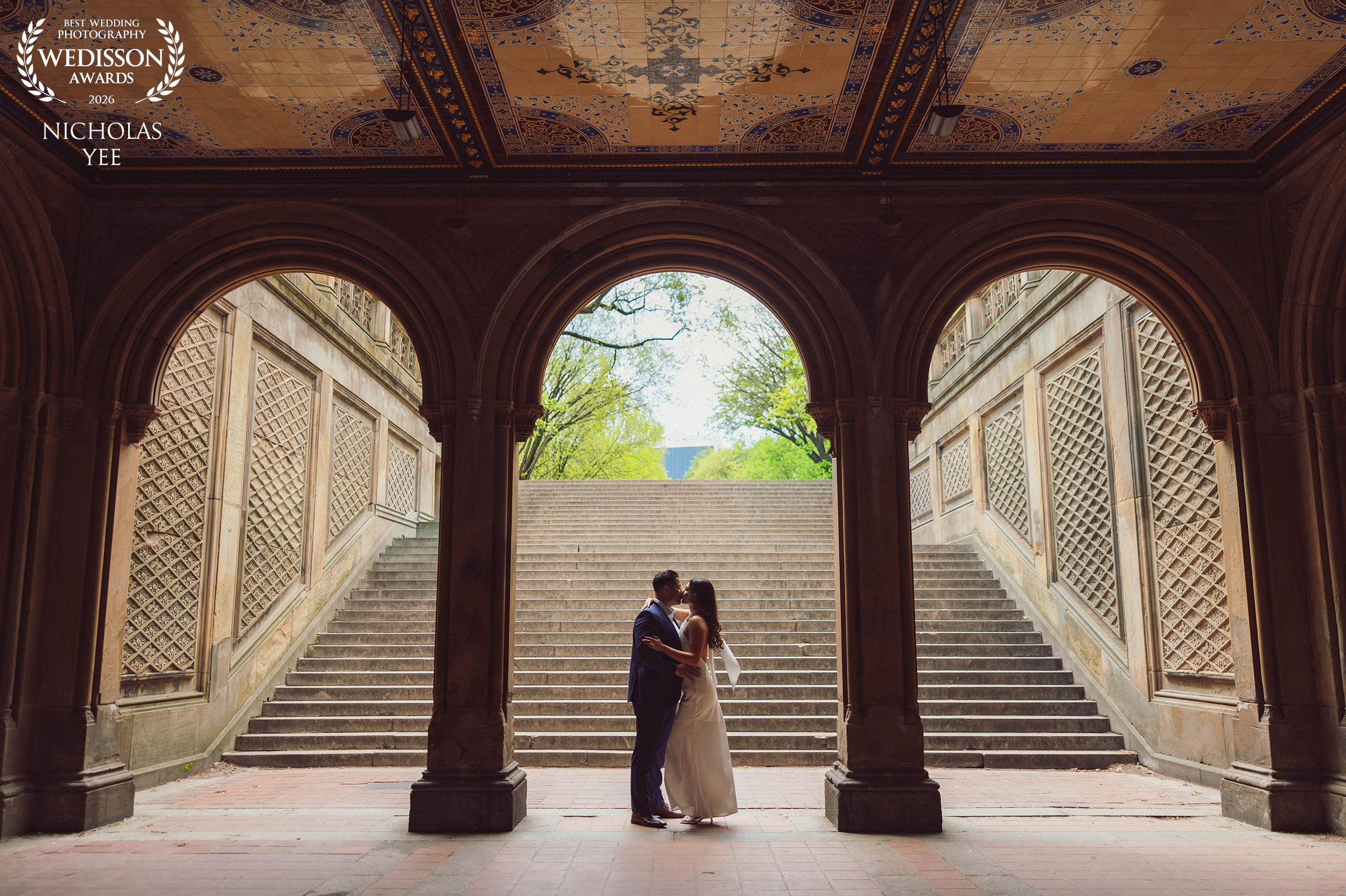 Beneath the Arches award-winning wedding photo by Nicholas Yee Photography from Wedisson Collection 134, featuring a couple kissing beneath ornate stone arches and a grand staircase.