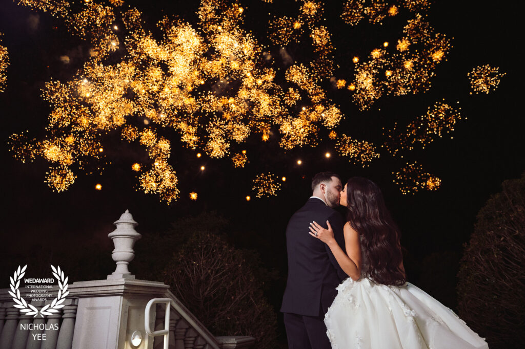 WedAward Collection #129 winning image by Nicholas Yee Photography featuring a bride and groom kissing at night beneath golden fireworks.