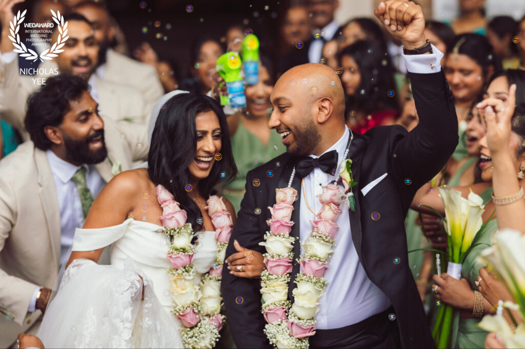 Award-winning wedding photo of a couple laughing with guests, floral garlands and bubbles by Nicholas Yee Photography.