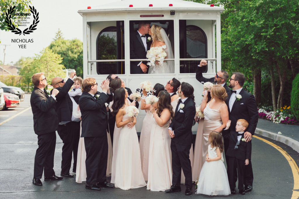 Bride and groom kissing aboard a white wedding trolley while their wedding party cheers below.