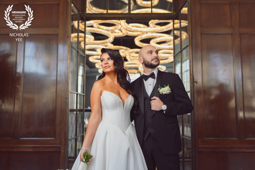 Bride in a structured white wedding gown and groom in a black tuxedo posing between wooden doors beneath sculptural circular lights.