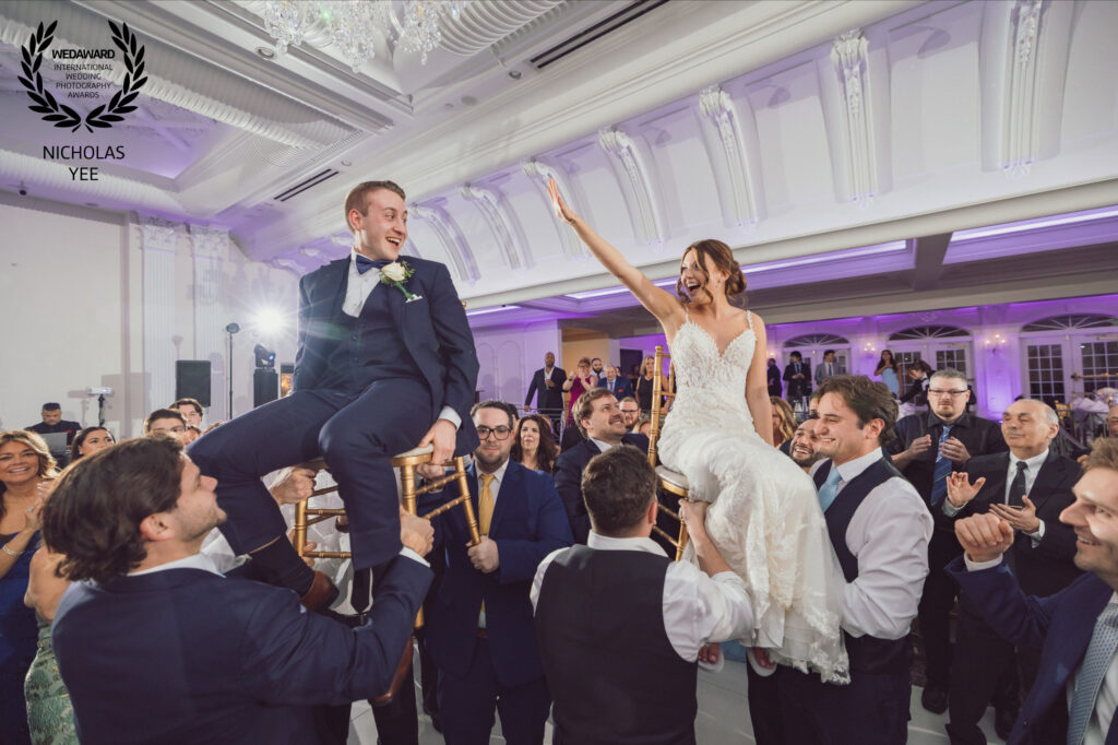 Bride and groom raised on chairs above cheering guests during an energetic wedding reception celebration.